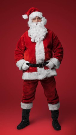 Foto de Jolly Santa Claus en traje rojo tradicional de pie con las manos en las caderas contra el fondo rojo - Imagen libre de derechos