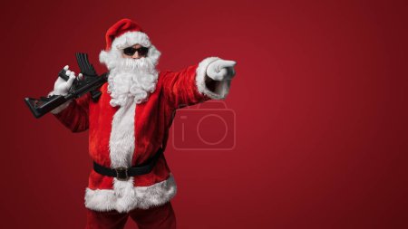 Téléchargez les photos : Un homme habillé comme le Père Noël, portant des lunettes de soleil noires élégantes, pose avec des mitrailleuses jouets sur un fond rouge audacieux - en image libre de droit