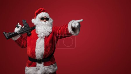 Téléchargez les photos : Un homme en costume de Père Noël, portant des lunettes de soleil noires, pose avec des pistolets jouets à la main sur un fond rouge - en image libre de droit