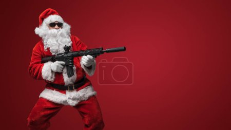 Photo for A man in a Santa Claus suit, wearing black sunglasses, poses with toy guns in hand against a red backdrop - Royalty Free Image