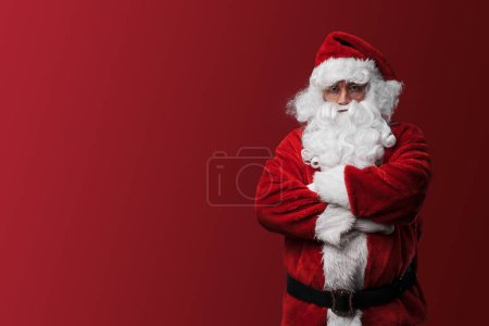 Téléchargez les photos : Sérieux Père Noël en costume rouge posant avec les bras croisés sur un fond rouge - en image libre de droit