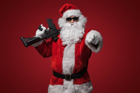 Téléchargez les photos : Un homme en costume de Père Noël, portant des lunettes de soleil noires, pose avec des pistolets jouets à la main sur un fond rouge - en image libre de droit