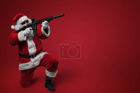Foto de Un hombre con un traje de Santa Claus, con gafas de sol negras, posa con armas de juguete en la mano contra un telón de fondo rojo - Imagen libre de derechos