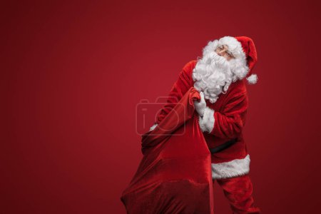 Téléchargez les photos : Père Noël portant un sac lourd, prêt à livrer des cadeaux de Noël - en image libre de droit