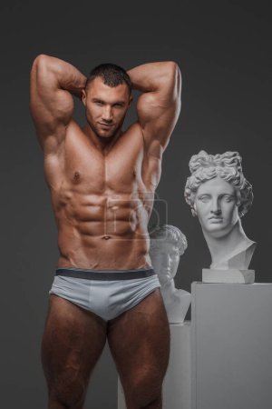 Handsome man exuding a rugged charm, with a flawless muscular chest, posing proudly beside ancient Greek statues against a gray backdrop