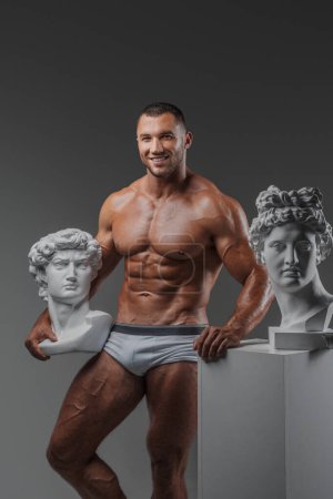 Téléchargez les photos : Portrait d'un homme souriant et robuste avec une apparence soignée, mettant en valeur son torse musculaire parfait nu tout en se tenant à côté de statues grecques antiques sur un gris - en image libre de droit
