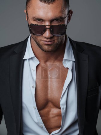 Foto de Modelo guapo en gafas de sol y una camisa abierta, revelando su torso muscular, sobre un fondo gris - Imagen libre de derechos