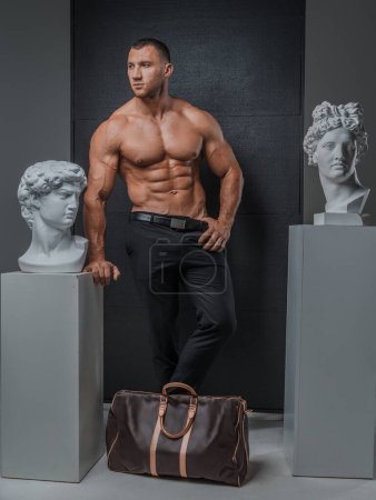 Mann mit gepflegtem Modelauftritt mit nacktem muskulösen Oberkörper, der mit einer luxuriösen High-Fashion-Tasche neben antiken griechischen Statuen auf grauem Hintergrund posiert