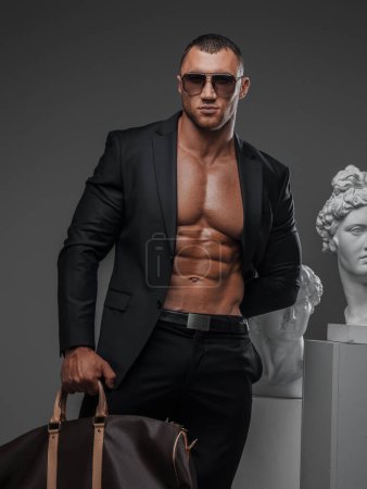Robuster Mann mit Sonnenbrille, offenem Anzug und muskulösem Oberkörper, der mit einer luxuriösen High-Fashion-Tasche neben antiken griechischen Statuen vor grauem Hintergrund posiert