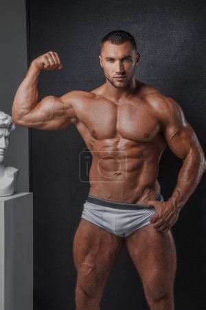 Foto de Retrato de un hombre resistente con una apariencia modelo bien arreglada, con calzoncillos blancos, y mostrando su torso muscular desnudo sobre un fondo gris - Imagen libre de derechos