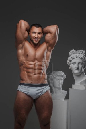 Foto de Retrato de un hombre sonriente y rugoso con una apariencia modelo bien arreglada, mostrando su torso muscular desnudo y perfecto mientras está de pie junto a estatuas griegas antiguas en un gris - Imagen libre de derechos