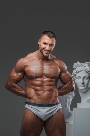 Téléchargez les photos : Portrait d'un homme souriant et robuste avec une apparence soignée, mettant en valeur son torse musculaire parfait nu tout en se tenant à côté de statues grecques antiques sur un gris - en image libre de droit