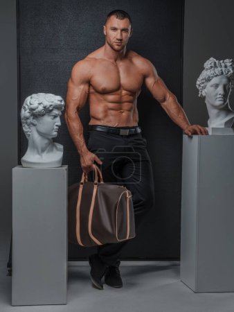Photo for Man with a well-groomed model appearance with a bare muscular torso, posing with a luxurious high-fashion bag next to ancient Greek statues on a gray background - Royalty Free Image