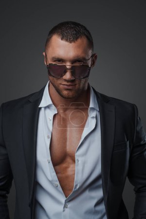 Handsome model in sunglasses and an open shirt, revealing his muscular torso, on a gray background