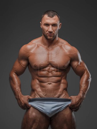Photo for Portrait of a rugged man with a well-groomed model appearance, wearing white briefs, and showcasing his bare muscular torso against a gray background - Royalty Free Image