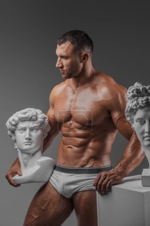 Photo for Handsome man exuding a rugged charm confidently showcasing his immaculate muscular physique while standing beside ancient Greek statues against a gray backdrop - Royalty Free Image