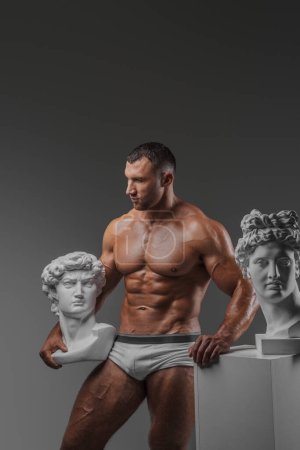 Téléchargez les photos : Bel homme exsudant un charme robuste mettant en valeur avec confiance son physique musculaire immaculé tout en se tenant à côté de statues grecques antiques sur un fond gris - en image libre de droit