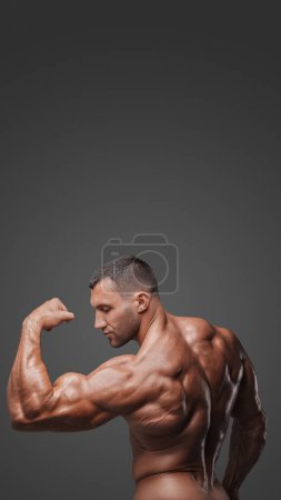 Foto de Vista desde atrás de un hombre musculoso con un torso desnudo, golpeando una pose, mostrando sus músculos tensos sobre un fondo gris - Imagen libre de derechos