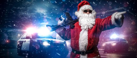 Téléchargez les photos : Le Père Noël, tenant une mitrailleuse, pointe son doigt quelque part, posant devant des voitures de police avec de nombreuses lumières et sirènes de police, au milieu d'une nuit de tempête enneigée dans la rue - en image libre de droit