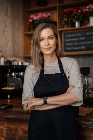 Photo for Smiling woman in apron standing confidently in a cozy cafe environment, with shelves of flowers in the background - Royalty Free Image
