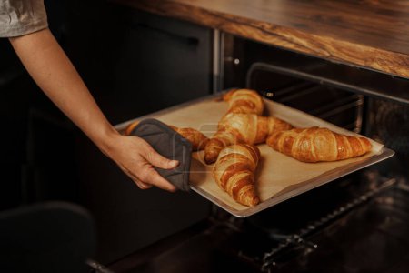 Foto de Una persona está quitando una bandeja de croissants marrón dorado de un horno, usando un guante protector del horno - Imagen libre de derechos