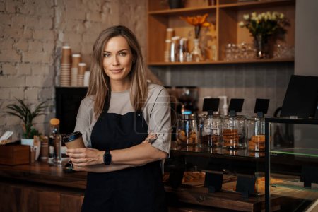 Photo for A portrait of a poised female barista holding a takeaway coffee cup, standing in her modern cafe - Royalty Free Image