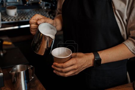 Foto de Primer plano de las manos de un barista vertiendo leche espumosa en una taza de café de papel, con una máquina de café espresso borrosa en el fondo - Imagen libre de derechos