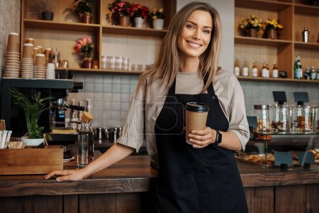 Téléchargez les photos : Portrait d'une serveuse tendue tenant une tasse de café à emporter, debout dans son café moderne - en image libre de droit
