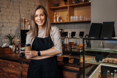 Téléchargez les photos : Portrait d'une serveuse tendue tenant une tasse de café à emporter, debout dans son café moderne - en image libre de droit