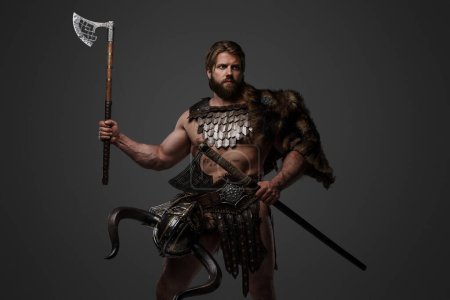 Foto de Un feroz guerrero vikingo barbudo de piel y armadura ligera, con un casco unido a su cinturón, sosteniendo un gran hacha de dos manos sobre un fondo gris - Imagen libre de derechos