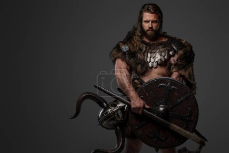 Foto de Un valiente guerrero vikingo con una majestuosa barba adornada con piel y armadura ligera, llevando un hacha y un escudo, de pie sobre un fondo gris - Imagen libre de derechos