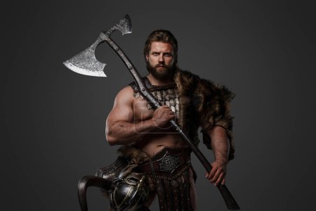 Ein grimmiger bärtiger Wikingerkrieger in Pelz und leichter Rüstung, mit einem Helm am Gürtel, der eine große beidhändige Axt auf grauem Hintergrund hält.
