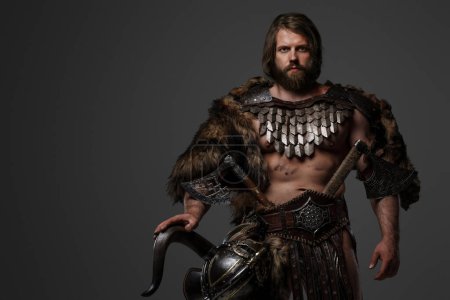 Téléchargez les photos : Un guerrier viking barbu féroce en fourrure et armure légère, avec un casque attaché à sa ceinture, tenant une grande hache à deux mains sur un fond gris - en image libre de droit
