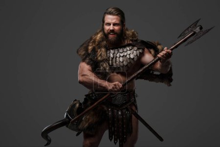 Foto de Un feroz guerrero vikingo barbudo vestido de piel y armadura ligera, sosteniendo un gran hacha de dos manos, gritando sobre un fondo gris - Imagen libre de derechos