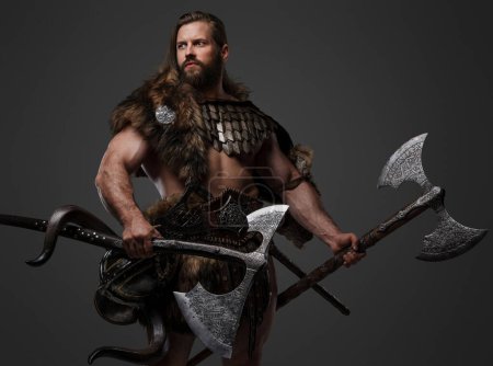 Téléchargez les photos : Un guerrier viking barbu féroce en fourrure et armure légère, avec un casque attaché à sa ceinture, tenant une grande hache à deux mains sur un fond gris - en image libre de droit