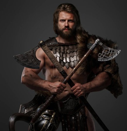 Foto de Un guerrero vikingo barbudo robusto vestido de piel y armadura ligera, con un casco unido a su cinturón y sosteniendo dos hachas sobre un fondo gris - Imagen libre de derechos