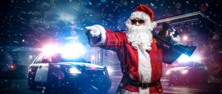Photo for Santa Claus, holding a machine gun, points his finger somewhere, posing in front of police cars with numerous police lights and sirens, amid a snowy stormy night on the street - Royalty Free Image