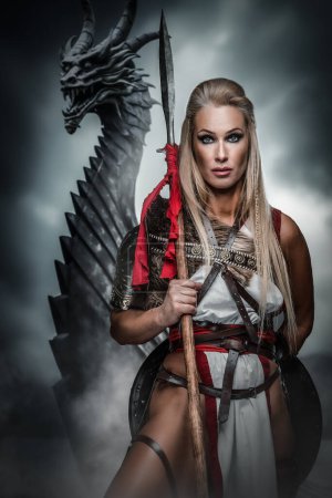 Téléchargez les photos : Une guerrière féroce se tient debout avec une lance devant un dragon menaçant, exsudant confiance et force dans un contexte orageux - en image libre de droit