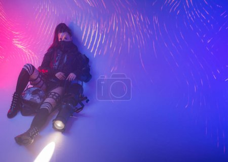 Photo for A female figure, dressed in a futuristic tactical black ensemble, reclines on the ground with a sizable projector against a backdrop of projected digital symbols - Royalty Free Image
