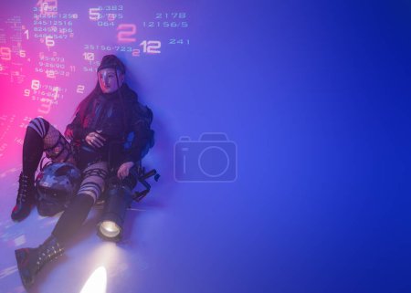 Foto de Una figura femenina, vestida con un conjunto negro táctico futurista, se reclina en el suelo con un proyector considerable sobre un fondo de símbolos digitales proyectados - Imagen libre de derechos