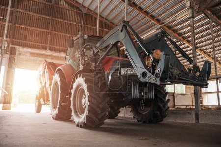 Téléchargez les photos : Un tracteur moderne rouge et noir équipé d'une chargeuse, garée à l'intérieur d'une grange en bois spacieuse - en image libre de droit