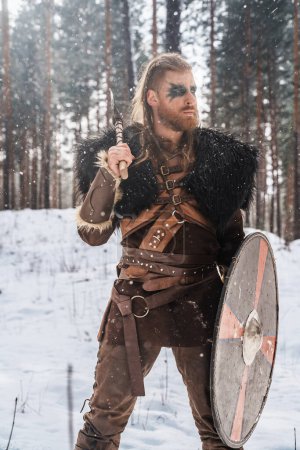 Photo for A fierce Viking warrior poised with an axe and shield in a snowy pine forest, depicting strength and historical Nordic culture - Royalty Free Image