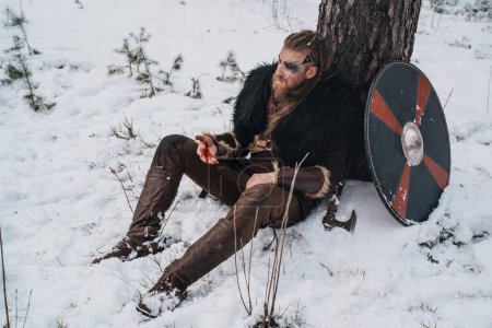 Foto de Un guerrero vikingo con pintura de guerra en la cara descansa contra un árbol en la nieve, apareciendo herido y exhausto - Imagen libre de derechos