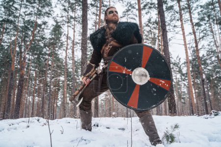 Photo for A man dressed as a Viking warrior stands with an axe and shield in a snowy forest, evoking historical Norse imagery - Royalty Free Image