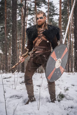Photo for A man dressed as a Viking warrior stands with an axe and shield in a snowy forest, evoking historical Norse imagery - Royalty Free Image