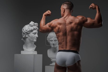 Foto de Hombre musculoso, vuelto a la cámara, flexiona sus músculos de la espalda bien definidos mientras está de pie en un estudio al lado de bustos griegos antiguos - Imagen libre de derechos