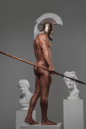 Photo for Muscular man stands sideways to the camera, posing nude with a spear in hand and a Greek helmet on his head while standing on a pedestal in a studio alongside ancient Greek busts - Royalty Free Image