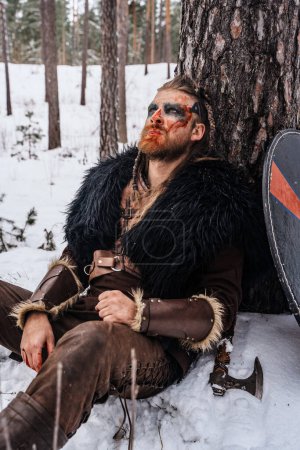 Foto de Un guerrero vikingo con pintura de guerra en la cara descansa contra un árbol en la nieve, apareciendo herido y exhausto - Imagen libre de derechos