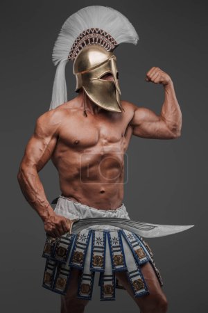 Photo for Portrait of an ancient Greek hoplite, posing in a studio with a bare muscular torso, wearing a helmet, and holding a gladius, set against a gray background - Royalty Free Image