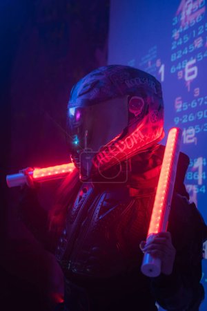 Foto de Una figura femenina, vestida con un conjunto negro táctico futurista y un casco de motocicleta, empuña dos lámparas de neón mientras se coloca delante de una pantalla de símbolos digitales proyectados - Imagen libre de derechos